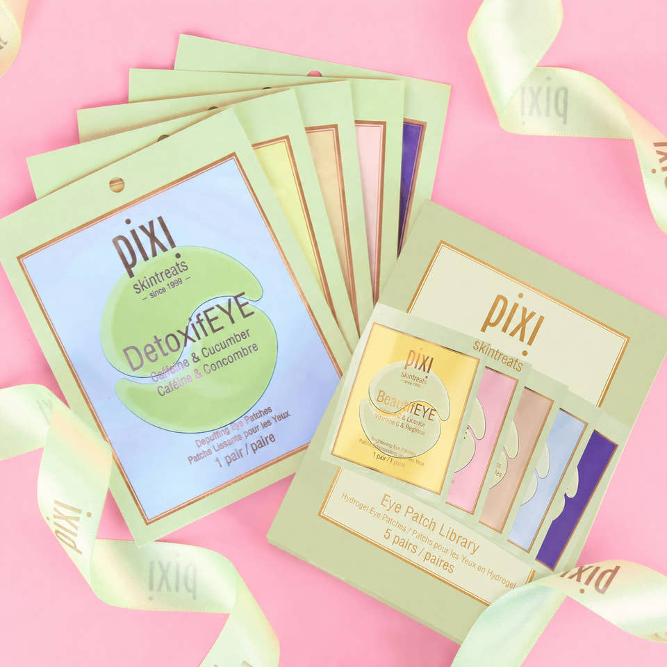 A styled flat-lay of Pixi Skinetreats eye patch sachets on a pink background with pale green ribbon accents. The image shows multiple individual sachet packs fanned out and a boxed 'Eye Patch Library' set, both branded Pixi. view 1