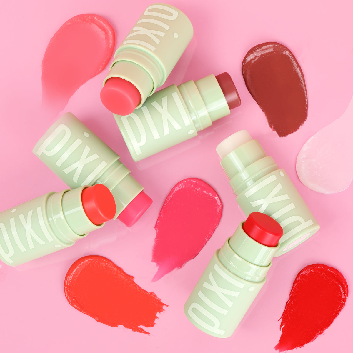 Four Pixi +HydraLip Treat sticks in pale Pixi green packaging arranged on a pink background with corresponding swatches in pink, coral, brown and red.