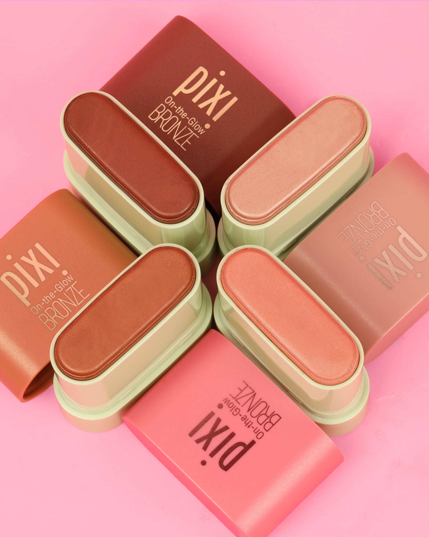 Pixi On-the-Glow Bronze makeup sticks arranged in a flat-lay pattern on a solid pink background, showing various warm-toned shades from deep brown to light peach.