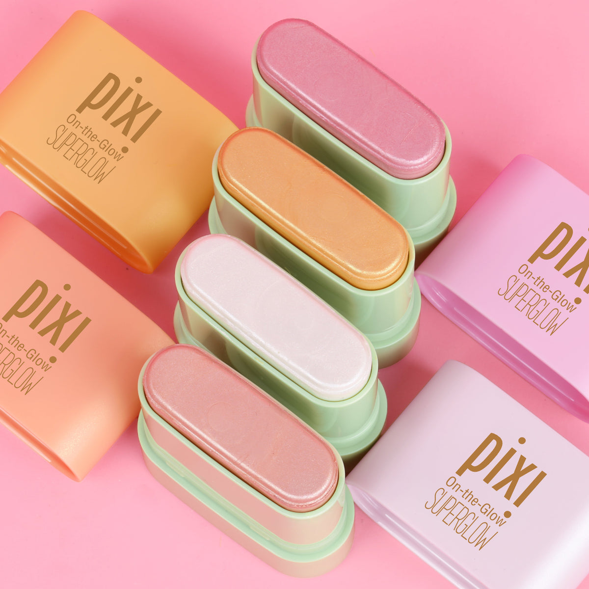 Six Pixi contour and highlight sticks in pastel shades of orange, peach, pink, mint green, white, and lavender, arranged diagonally on a solid pink background.