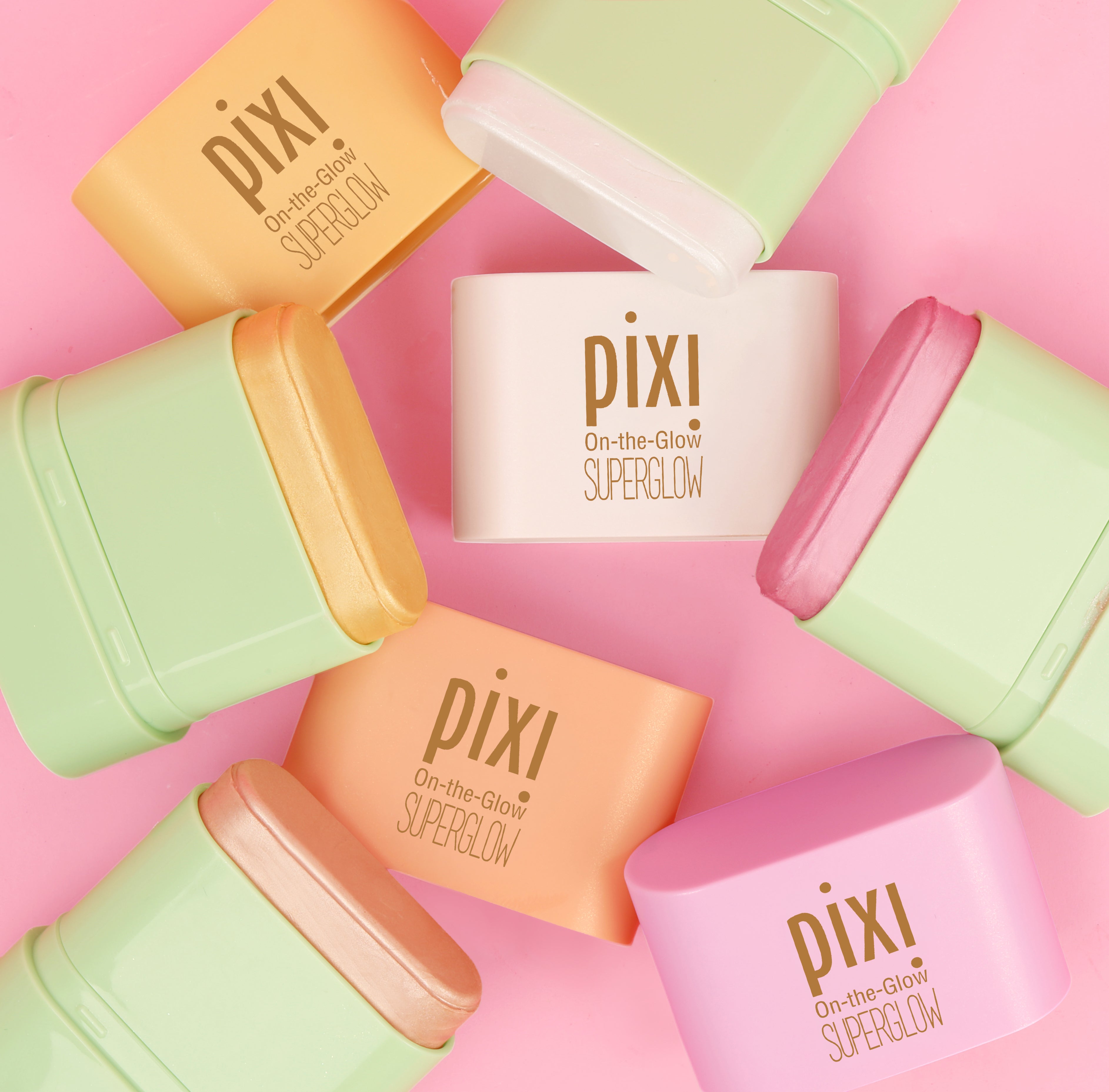 Pixi On-the-Glow SuperGlow cream products in pastel shades arranged in a circular flat-lay pattern on a solid pink background.
