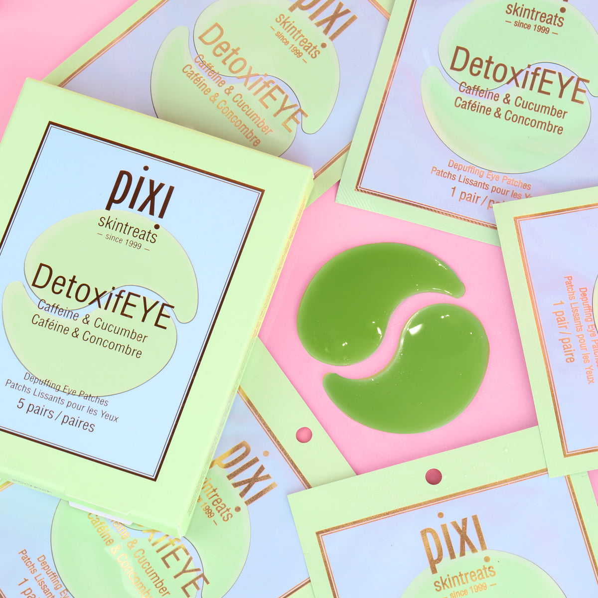 Several Pixi Skintreats DetoxiEYE caffeine and cucumber eye patch packets are arranged on a pink background, with one packet opened to show a green crescent-shaped eye patch in the center.