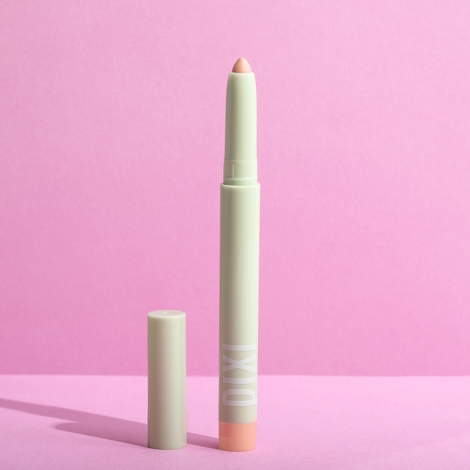 Pixi CC Crayon concealer pencil in a pale mint green shade with a peach-colored tip, shown next to its cap on a pink background. view 1
